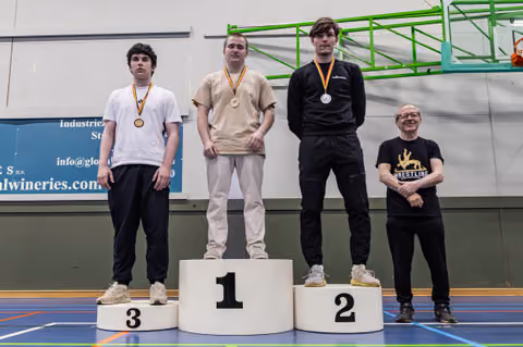 Three young wrestlers stand on a podium (1st, 2nd, 3rd), wearing medals, with a coach smiling beside them in a sports hall.
