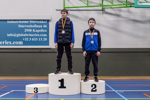 Two young boys stand on a podium, first and second place, wearing medals in a Belgian indoor sports hall.