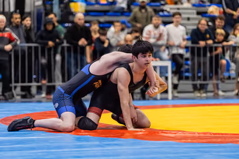 Young wrestler in black singlet holds position on mat, looking focused, as opponent in blue applies pressure from behind.