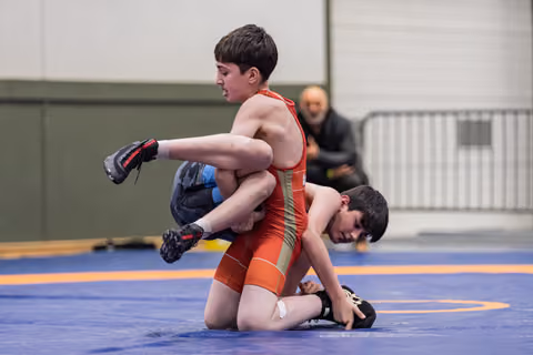 Boy in red singlet lifts opponent off the mat during an intense youth wrestling bout on a blue competition mat.
