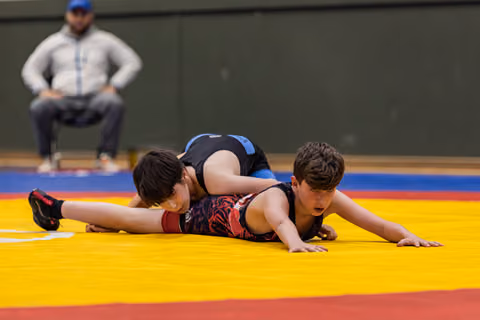 Two young boys wrestling on a yellow mat, one pinning the other down, coach watching intently in the background.