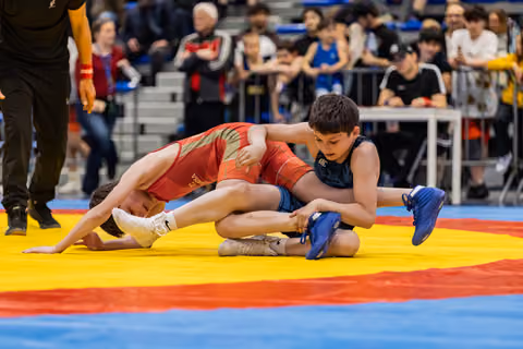 Two youth wrestlers battle on a colorful mat, one in red being controlled by opponent in blue grabbing his leg.