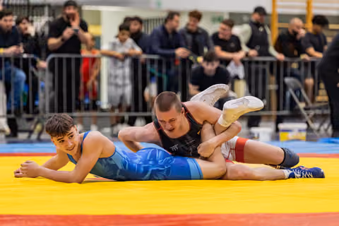 Young wrestler in blue singlet smiles while being pinned by opponent in black, on a yellow and red mat.