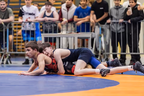 Young wrestler in red struggles on all fours as opponent in navy controls him from above on orange and blue mat.