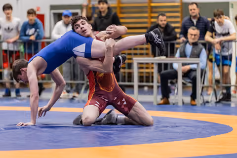 Wrestler in red singlet lifts opponent's leg aggressively on blue mat, crowd watching intently in background.