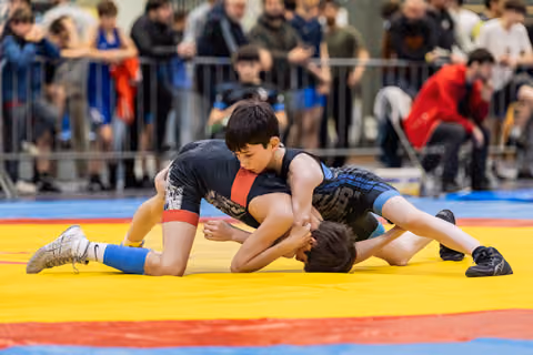 Young wrestler dominates opponent on yellow mat, pressing down with focused intensity during indoor tournament.
