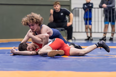 Curly-haired wrestler in navy singlet pins opponent in red to the mat, both showing intense strain during match.