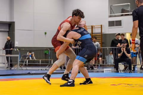 Two wrestlers locked in a fierce clinch on a yellow mat, red singlet athlete driving forward aggressively in an indoor sports hall.