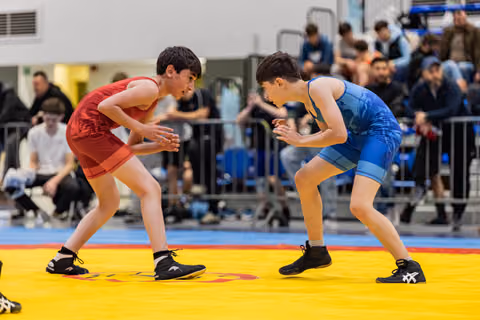 Two young wrestlers in red and blue singlets face off in a crouched ready stance on a yellow mat, crowd watching.