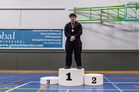 Young male athlete stands proudly on the first-place podium, wearing a gold medal, in an indoor sports hall.