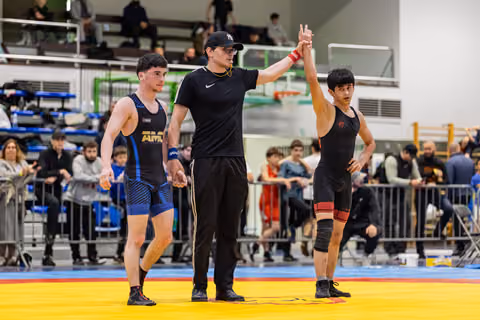Referee raises the hand of the winner in red singlet as the defeated wrestler in blue stands nearby on the mat.