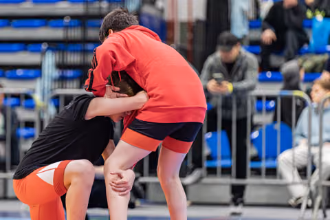 Two young wrestlers locked in a tight clinch on the mat, both leaning forward with intense focus during an indoor event.