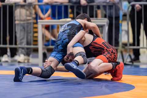 Two young wrestlers compete intensely on a blue mat, one in blue dominating from above, the other in red defending below.