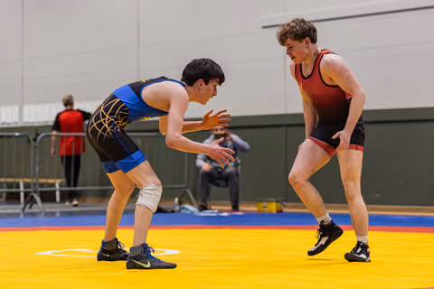 Two young wrestlers in blue and red singlets square off in a crouched stance, focused and ready to engage on a yellow mat.