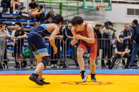 Two young male wrestlers in navy and red singlets square off in a crouched stance, focused and tense, on a yellow mat.