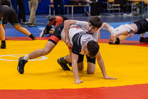 Young wrestler dominates opponent from above on yellow mat, both focused and intense during indoor tournament.