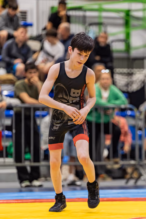 Young wrestler in black singlet moves with intense focus on colorful competition mat, crowd watching behind barriers.