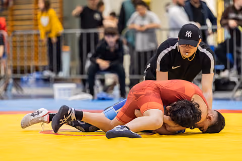 Referee in NY cap watches closely as wrestler in red singlet pins opponent face-down on yellow mat.