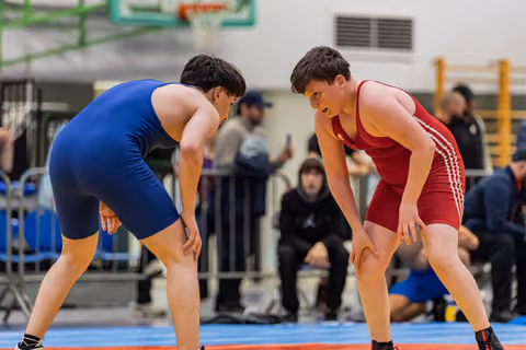 Two young wrestlers in blue and red singlets crouch low, locked in intense eye contact before engaging on the mat.