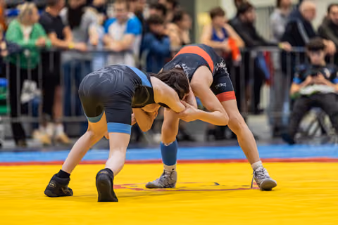 Two young wrestlers locked in a grappling clinch, bent forward in intense focus on a yellow competition mat.
