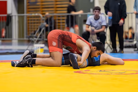 Wrestler in red singlet dominates opponent in blue, pressing them down on the yellow competition mat.