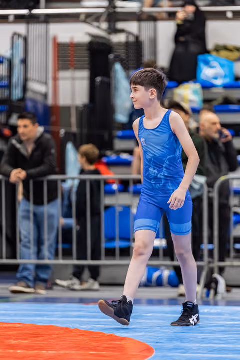Young boy in blue singlet walks confidently across the mat during a wrestling event, spectators visible in background.