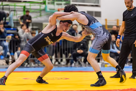 Two young wrestlers battle in a fierce clinch on a yellow mat, referee watching closely nearby.