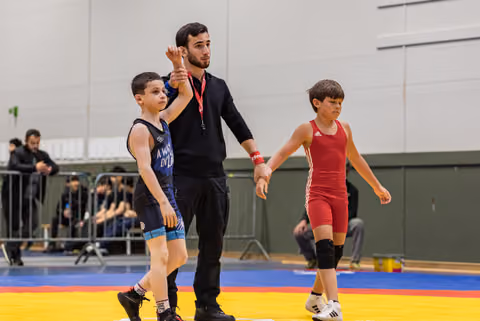 Referee raises the arm of a young wrestler in blue singlet declaring victory, as defeated opponent in red walks away.