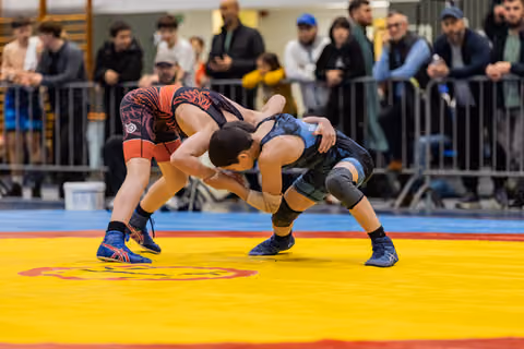 Two young wrestlers lock in an intense clinch on a yellow mat, crowd watching closely behind metal barriers.