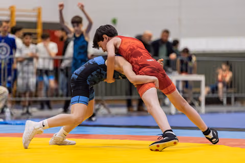 Two young wrestlers locked in a tight clinch on a yellow mat, crowd cheering energetically in the background.
