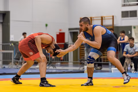 Two wrestlers engage on a yellow mat; blue-singlet athlete lunges forward, gripping opponent bent low in red.