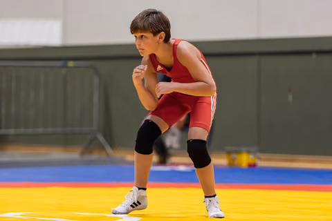Young wrestler in red Adidas singlet crouches in fighting stance, focused and intense, on colorful competition mat.