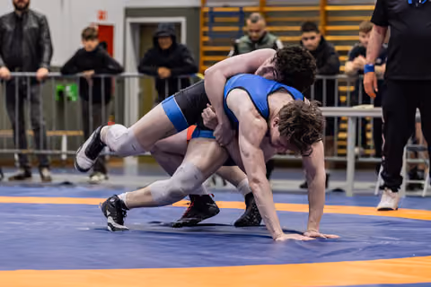 Two teenage wrestlers locked in a fierce grapple, one in blue singlet driving forward on a blue and orange mat.