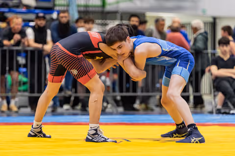 Two young wrestlers lock in a tense clinch on a yellow mat, both leaning forward with focused intensity before spectators.