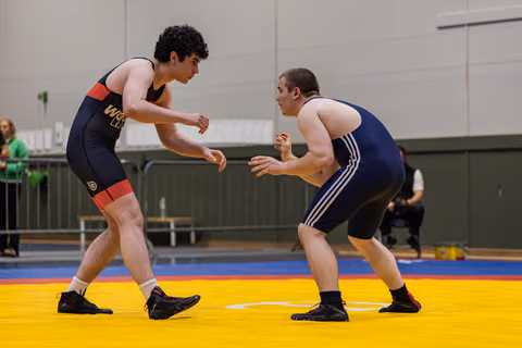 Two male wrestlers in fighting stance, circling each other intensely on a yellow mat in an indoor sports hall.