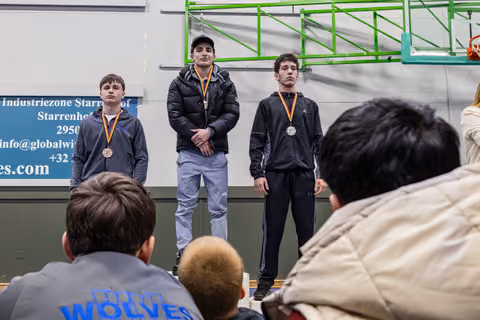 Three teenage wrestlers stand on a podium wearing medals, expressions serious and composed, in an indoor gymnasium.
