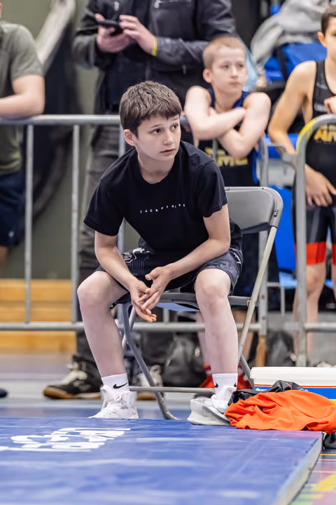 Young boy in black shirt sits tensely on a folding chair courtside, hands clasped, watching the match with focused anticipation.