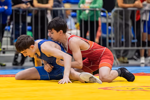 Teen in red singlet pins opponent in blue to yellow mat, both showing intense focus during match.