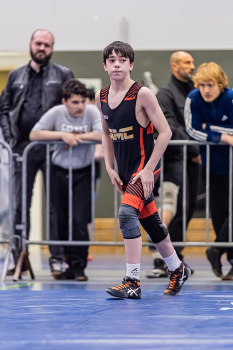 Young wrestler in black and red singlet stands alert on blue mat, focused expression, coaches watching behind barriers.