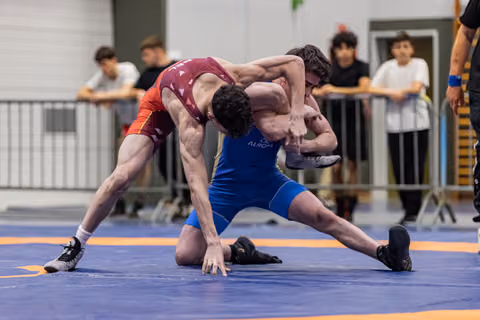Two young wrestlers battle for control, one in red driving forward, one in blue defending on the mat.