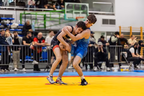 Two young wrestlers locked in an intense clinch on a yellow mat, crowd watching in a packed indoor sports hall.
