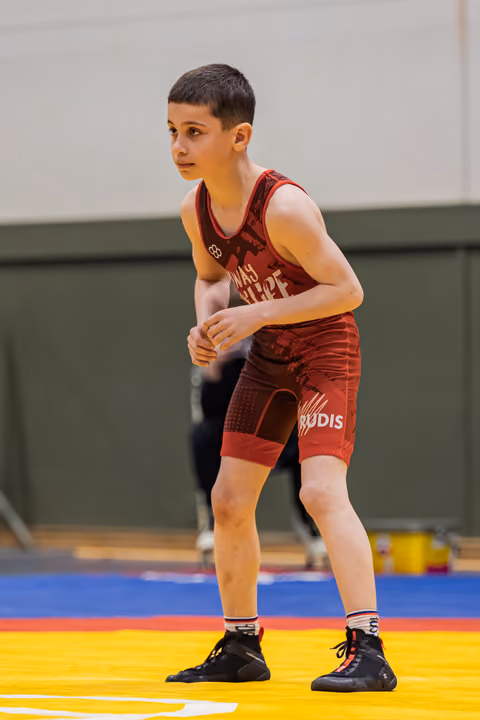 Young male wrestler in red singlet holds a focused defensive stance on a colorful mat, ready to compete.