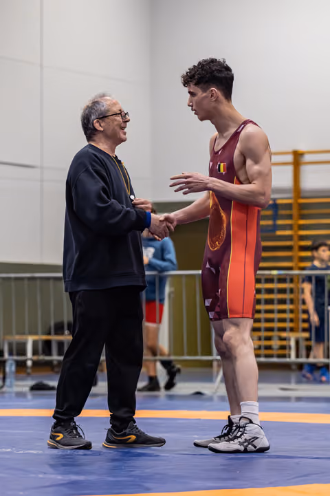 Older coach smiling warmly shakes hands with young male wrestler in maroon singlet on blue competition mat