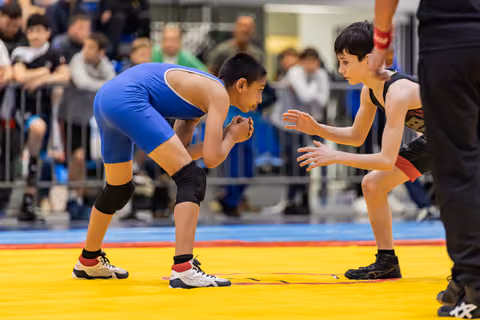Two young wrestlers in blue and black singlets face off on a yellow mat, focused and tense, crowd watching behind.