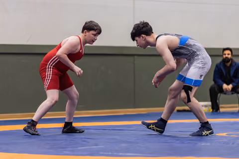 Two young wrestlers in red and grey singlets square off intensely on a blue competition mat, referee watching behind.