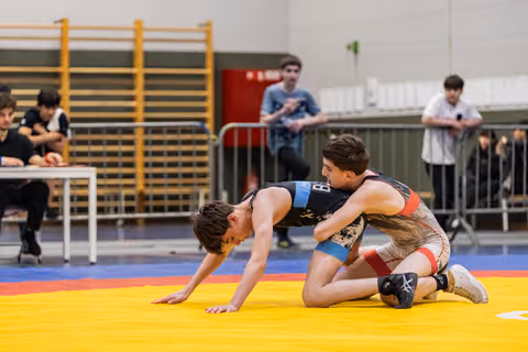Teen wrestler pinned down on all fours while opponent applies upper body control on a colorful mat.