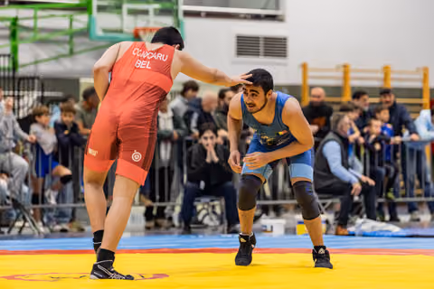 Two wrestlers in red and blue singlets face off on a yellow mat, crowd watching intently in the background.