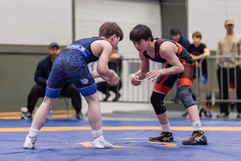Two young wrestlers in blue and red singlets crouch in tense standoff on blue mat, spectators watching behind barriers.