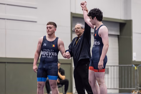 Referee raises the fist of a victorious wrestler in a navy Wolves singlet, while his defeated opponent stands nearby.