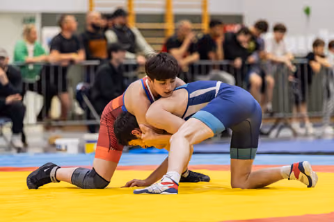Wrestler in blue singlet dominates opponent in red, both on knees grappling intensely on yellow mat, crowd watching.